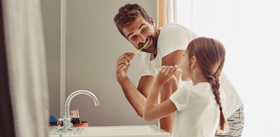 Dad & young daughter in white T-shirts leaning over the sink brushing their teeth together